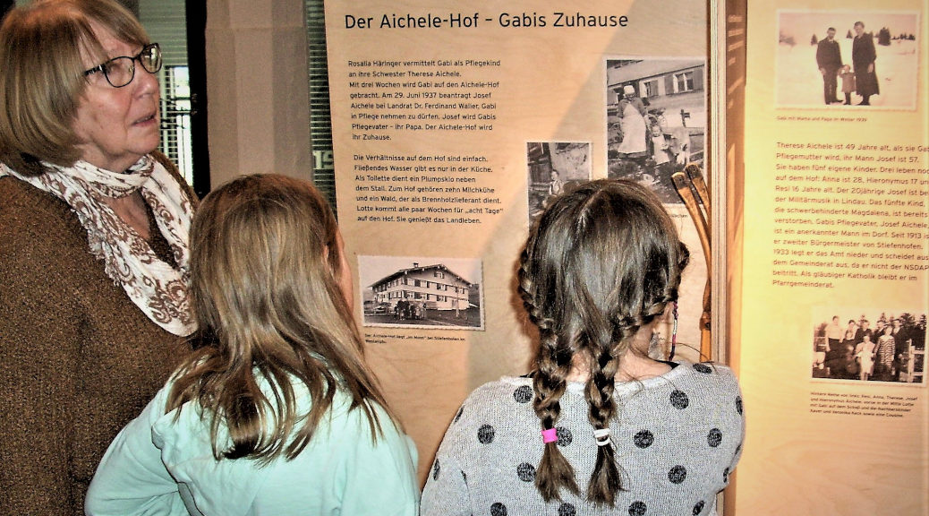 Rita Landsbeck mit zwei Enkelinnen an einer Tafel der Wanderausstellung Geliegte Gabi im Hutmuseum Lindenberg.
